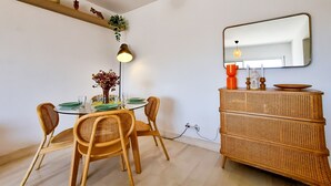 A dining area with a round table, four wicker chairs, a wicker sideboard, a wall-mounted mirror, and a hanging light fixture.
