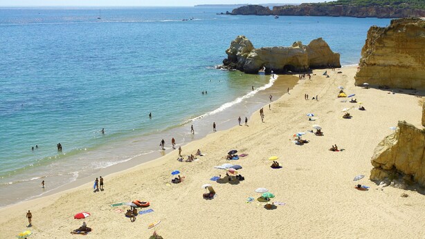 A beach with clear blue water, rocky cliffs, and people sunbathing.