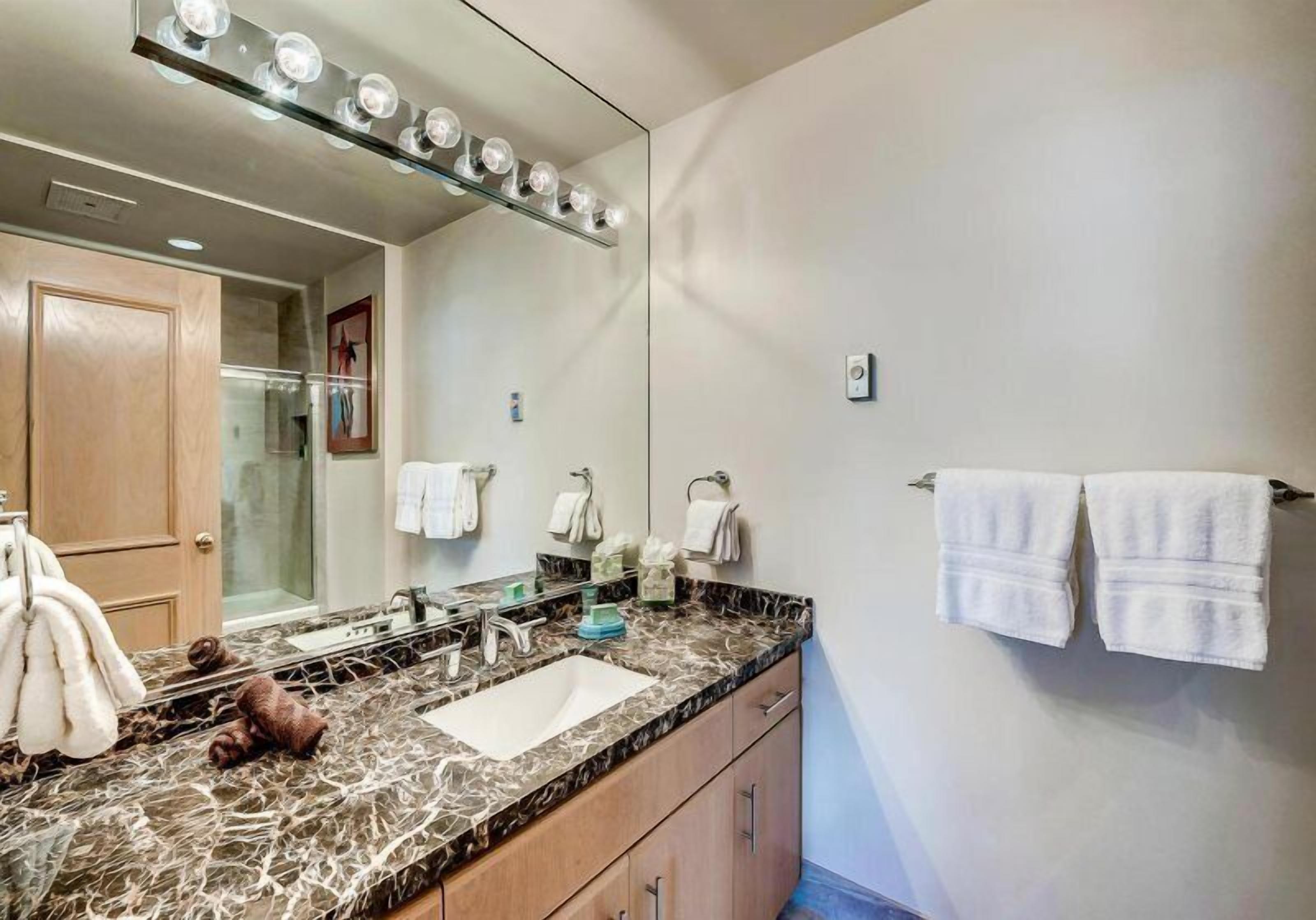 A bathroom with a marble countertop, a sink, a mirror with lights, and towels on a rack.