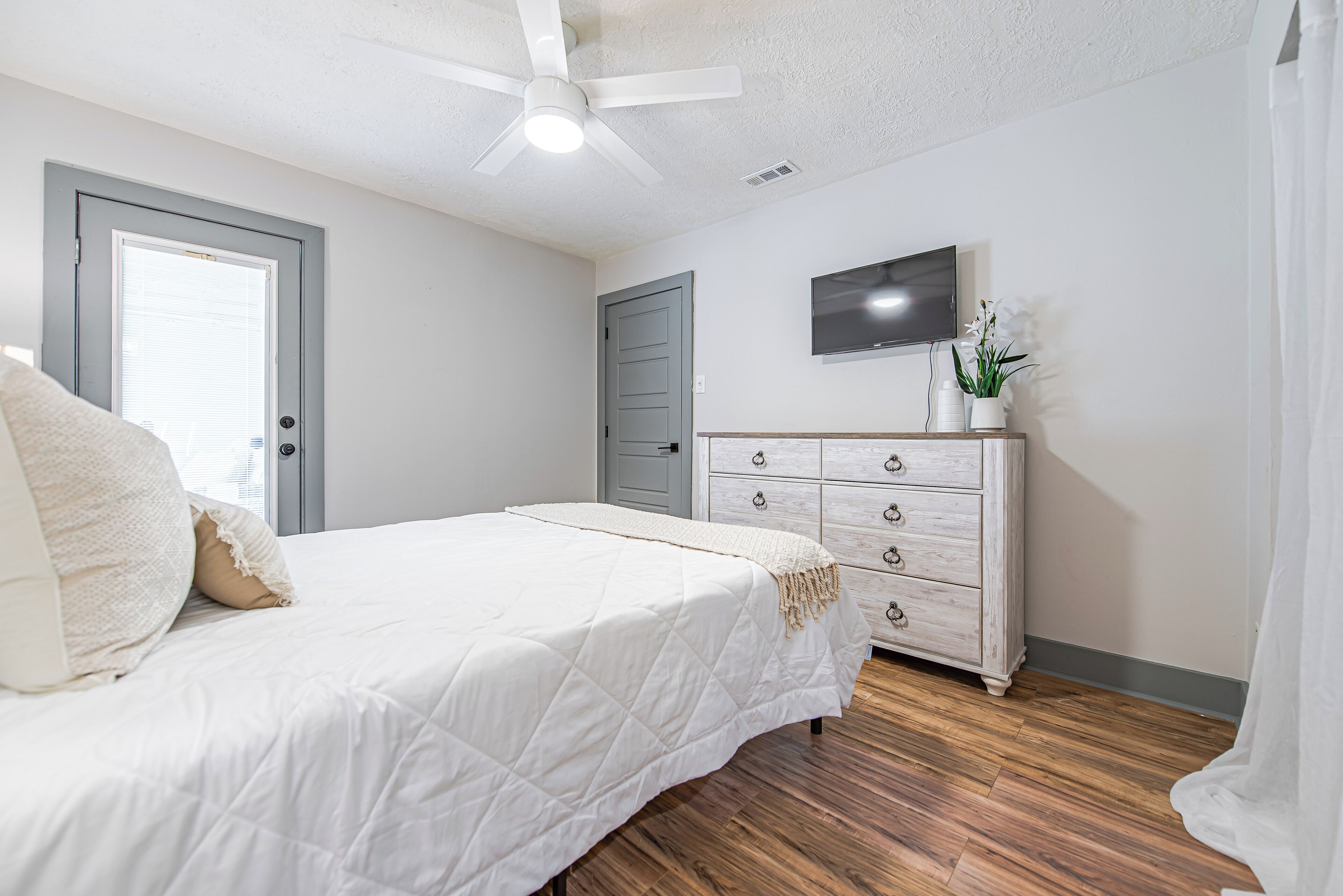Une chambre à coucher avec un lit, une commode, un ventilateur de plafond et un téléviseur fixé au mur.