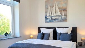 Una camera da letto con un letto grande, un comodino, una finestra vista verde e un quadro con barche a vela.