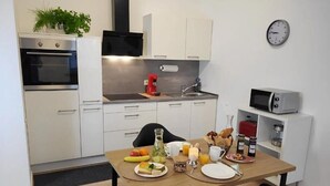 Una cucina moderna con mobili bianchi, forno integrato, macchina del caffè e microonde. È presente un tavolo da pranzo con prodotti per la colazione e un orologio a parete.