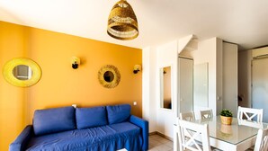 A room with a blue sofa, a glass dining table, white chairs, and a yellow accent wall. - La Cabre d'Or - Duplex climatisé (Cabriès)
