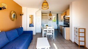 A compact living space with a blue sofa, a dining table, white chairs, a kitchen area with wooden cabinets, and a hanging rattan light fixture. - La Cabre d'Or - Duplex climatisé (Cabriès)