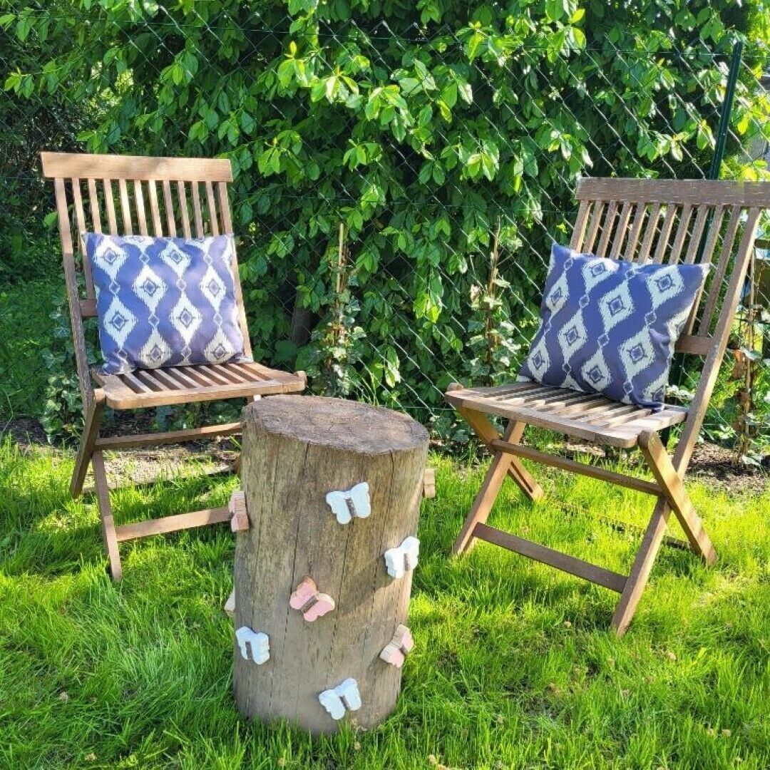 Two wooden chairs with patterned cushions, a wooden stump with butterfly decorations, and a lush green hedge in the background.
