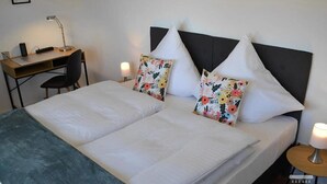 A double bed with white linens, two floral pillows, a desk with a lamp, and a chair. - Gästehaus am Bachlauf - Losheim am See 
Wohnung 6 – Weitblick (Losheim am See)