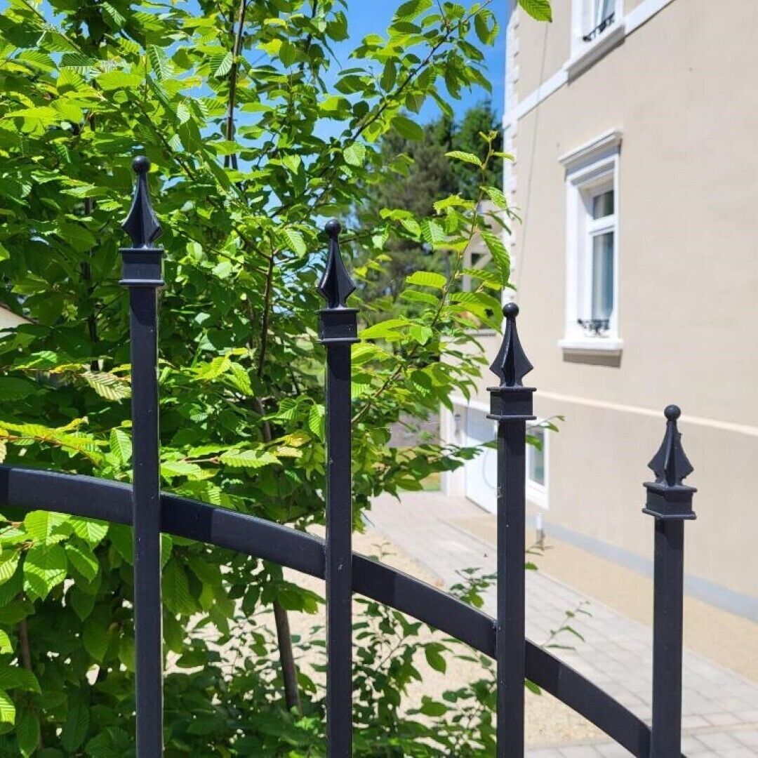 Ett svart metallstaket med spetsiga toppar, ett träd med gröna löv och en beige byggnad med ett fönster.
