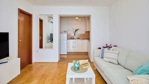 Un salon moderne avec un canapé, une table basse et un téléviseur. On aperçoit au fond une cuisine équipée de placards et d’un réfrigérateur.