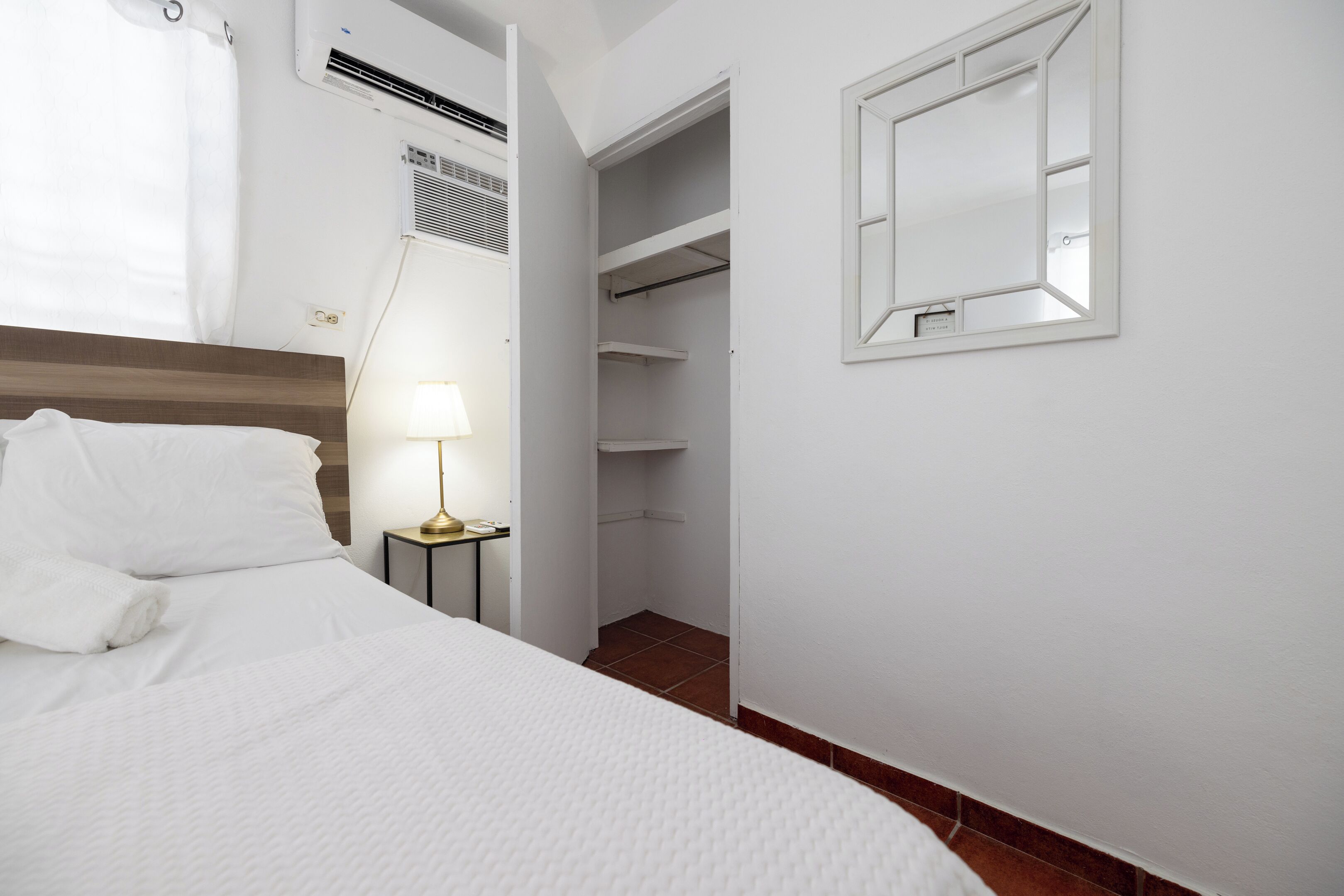 Una cama bien hecha con sábanas blancas, una lámpara de noche, un equipo de aire acondicionado, un armario blanco con puertas abiertas y una mesita con lámpara.