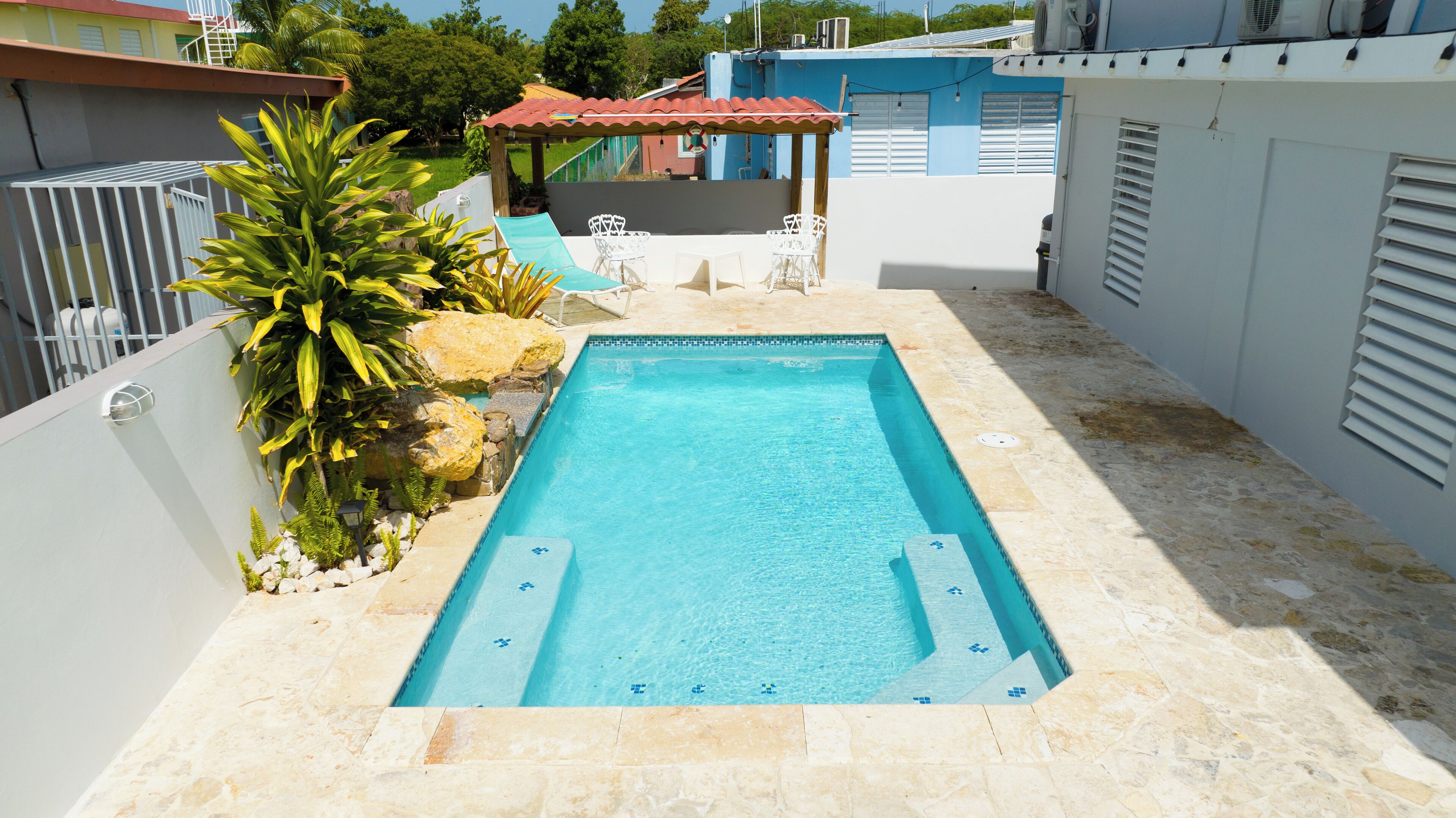 Una piscina rectangular con agua azul cristalina, rodeada por una terraza de piedra. Hay sillas blancas y una mesita bajo una zona con techo rojo.