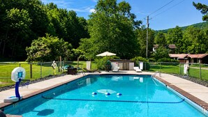 Una piscina rectangular con borde de azulejos, aro de baloncesto y una pequeña zona cubierta con asientos.