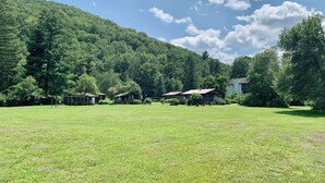 Entorno rural con un campo cubierto de hierba, algunos edificios y una colina boscosa.