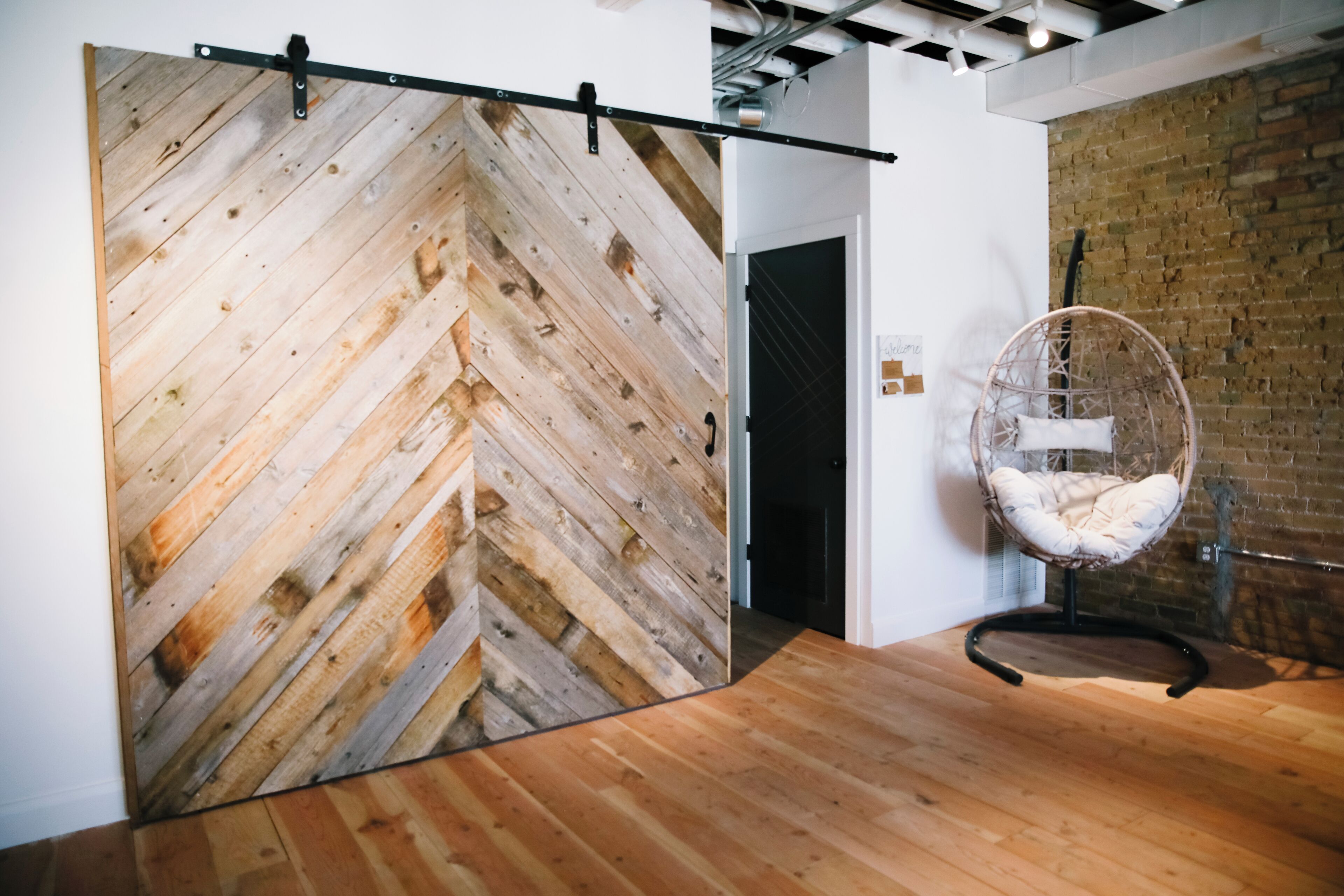 Quarto com piso de madeira em padrão herringbone, porta preta e cadeira de vime.