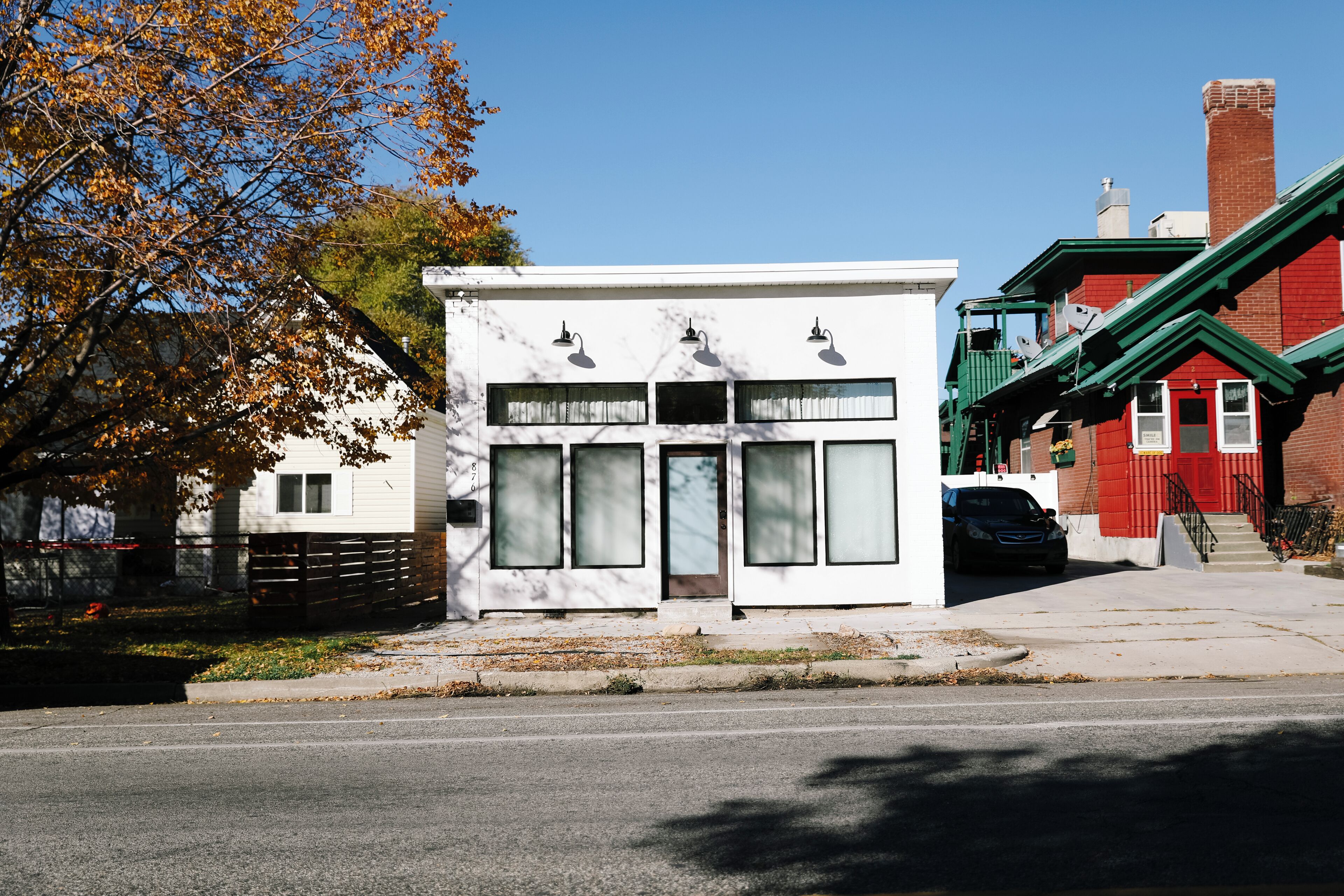 A white building with large windows and a door, a black car parked in front, and a red building with a green roof in the background.