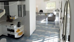 Eine moderne Küche mit weißen Schränken, einem schwarzen Wasserkocher und einem gefliesten Boden. Es gibt einen Essbereich mit einer Couch und einem Tisch.