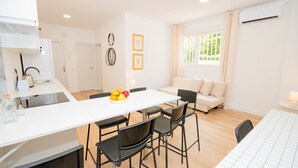 Cocina moderna con encimera blanca, una mesa de comedor con sillas y un frutero. Hay un sofá y una ventana con cortinas en la sala de estar.