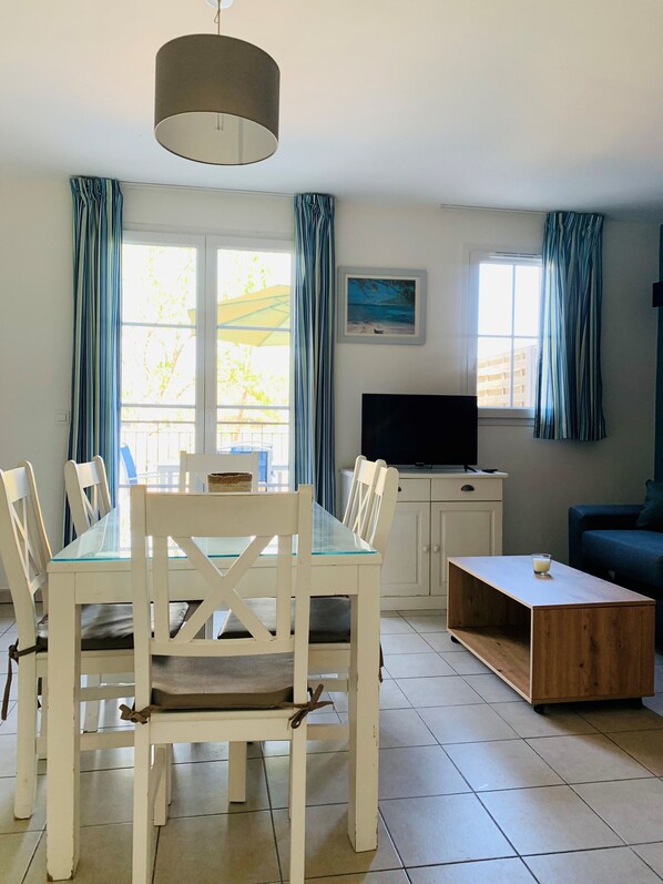 Un área de comedor con una mesa y sillas, un televisor sobre un mueble bajo, y una ventana con cortinas.