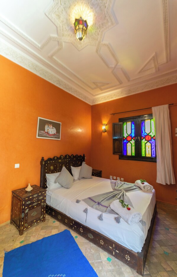 A bedroom with a bed, a nightstand, a wall-mounted light, a stained glass window, and a painting. - Riad Full Moon (Marrakech)