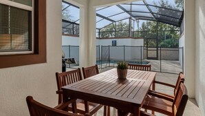 Un patio exterior con mesa y sillas de madera, piscina y una zona cubierta con vistas a árboles.