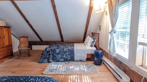 A bedroom with a sloped ceiling, wooden beams, a bed with patterned bedding, a small chair, a blue rug, and a window with blinds.