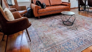 A spacious living room with a brown leather sofa, a patterned rug, and wooden beams on the ceiling.