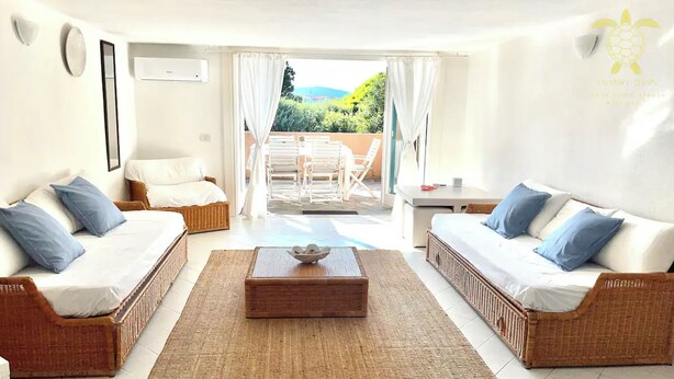 Una habitación luminosa y bien iluminada con dos sofás de mimbre, una mesita de centro de madera y vistas a un patio con mobiliario exterior.