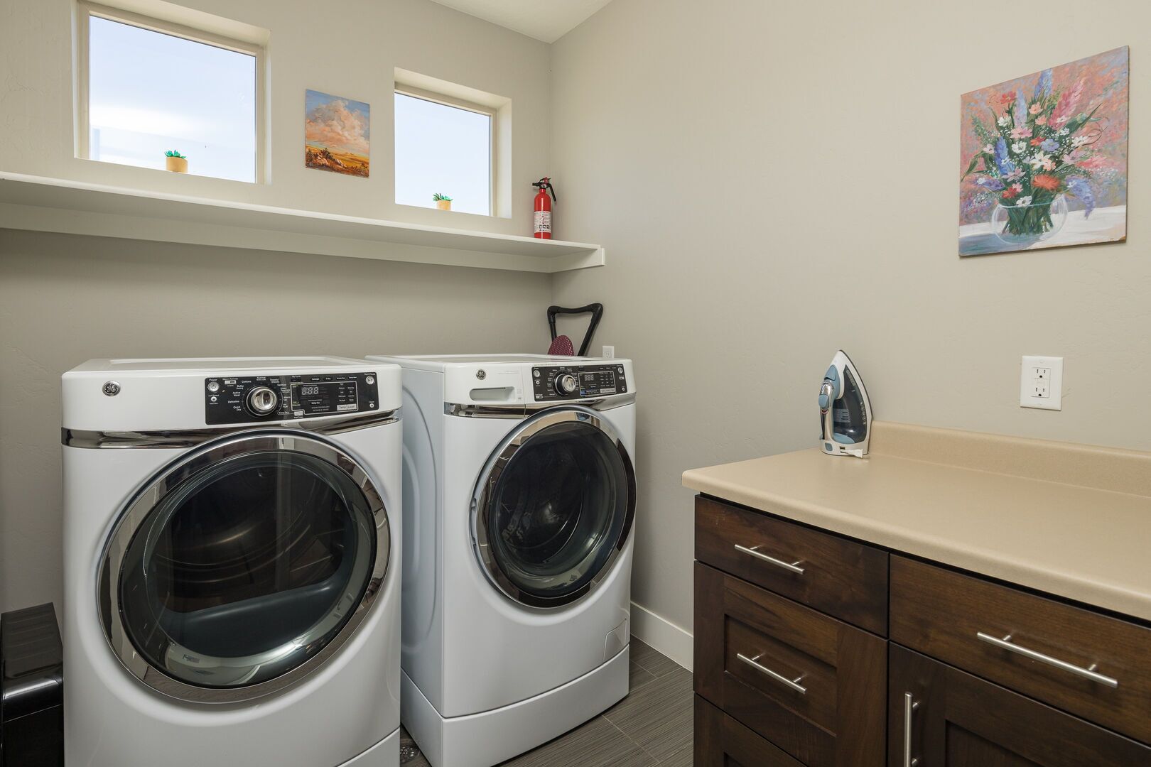 A laundry room with two front-loading washing machines, a countertop with a sink, and a painting on the wall.