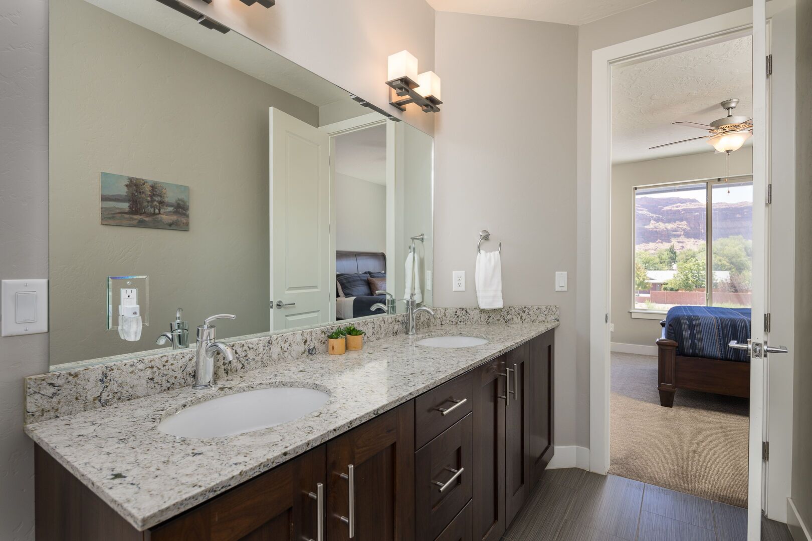 A bathroom with two sinks, a large mirror, and a door leading to a bedroom.