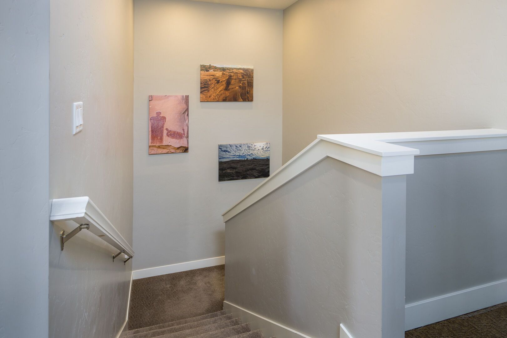 Un escalier avec une rampe, un interrupteur fixé au mur et trois œuvres d’art encadrées.