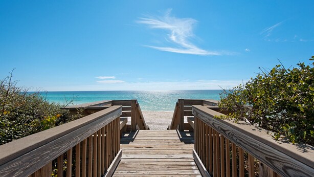 Ein Holzsteg führt zu einem Strand mit klarem, blauem Wasser und strahlend blauem Himmel.