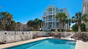 A pool area with a two-story building in the background, palm trees, and a white fence.