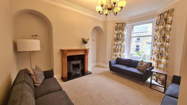 A living room with a fireplace, a sofa, a lamp, and a view of the outside through large windows. - Brylie House, Large Central Family Home (Skipton)