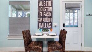 A dining area with a table set for two, two wicker chairs, a wall-mounted sign listing cities, and a window with blinds. - Romantic~Butterfly Cttg~Central Loc, Firepit ~Oaks (San Antonio)