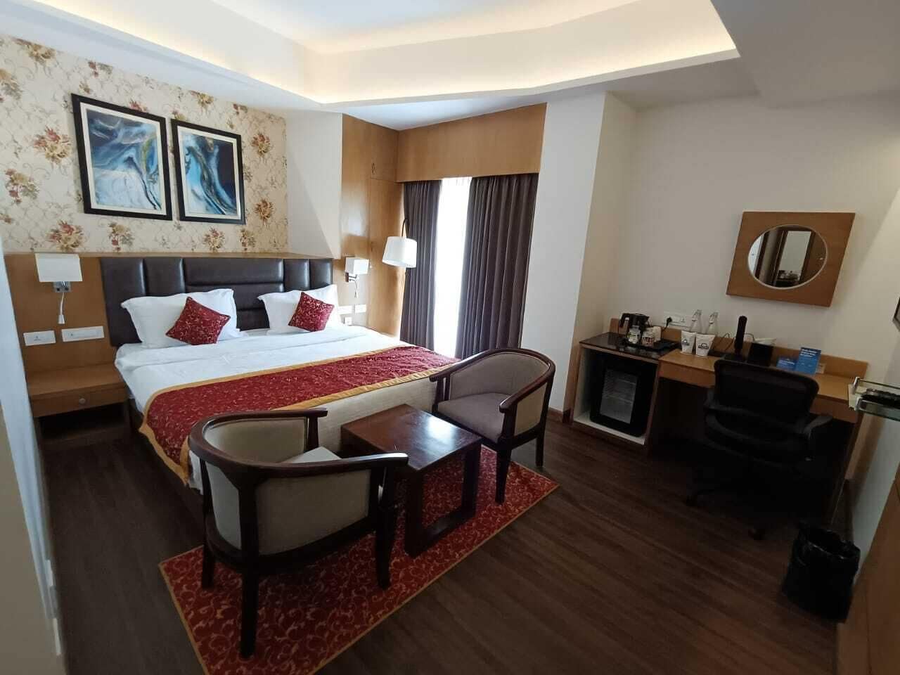 A hotel room with a large bed, a desk with a chair, a small table, a coffee machine, and a TV.