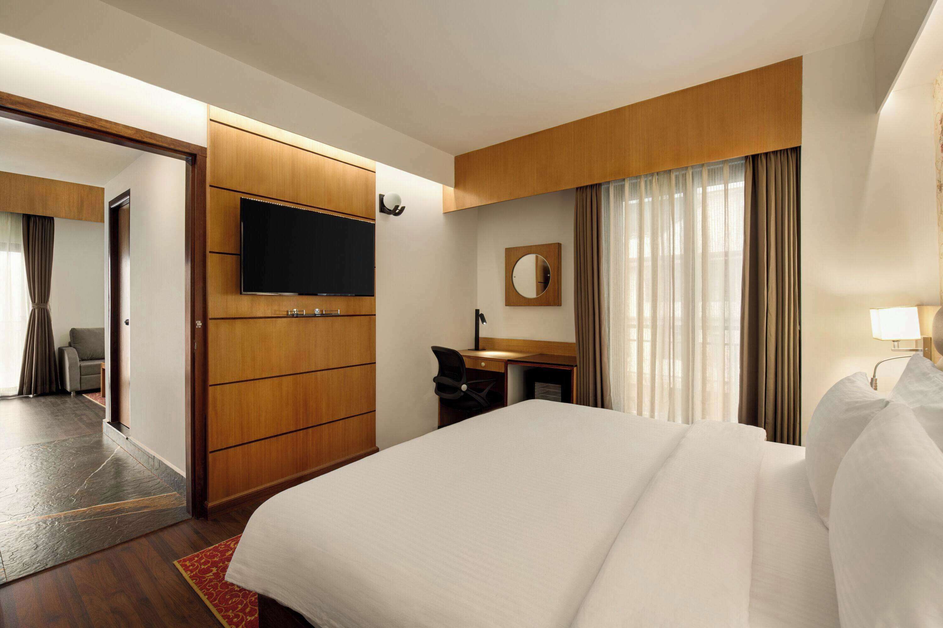 A hotel room with a large bed, a desk with a chair, a flat-screen TV, a mirror, and a window with curtains.