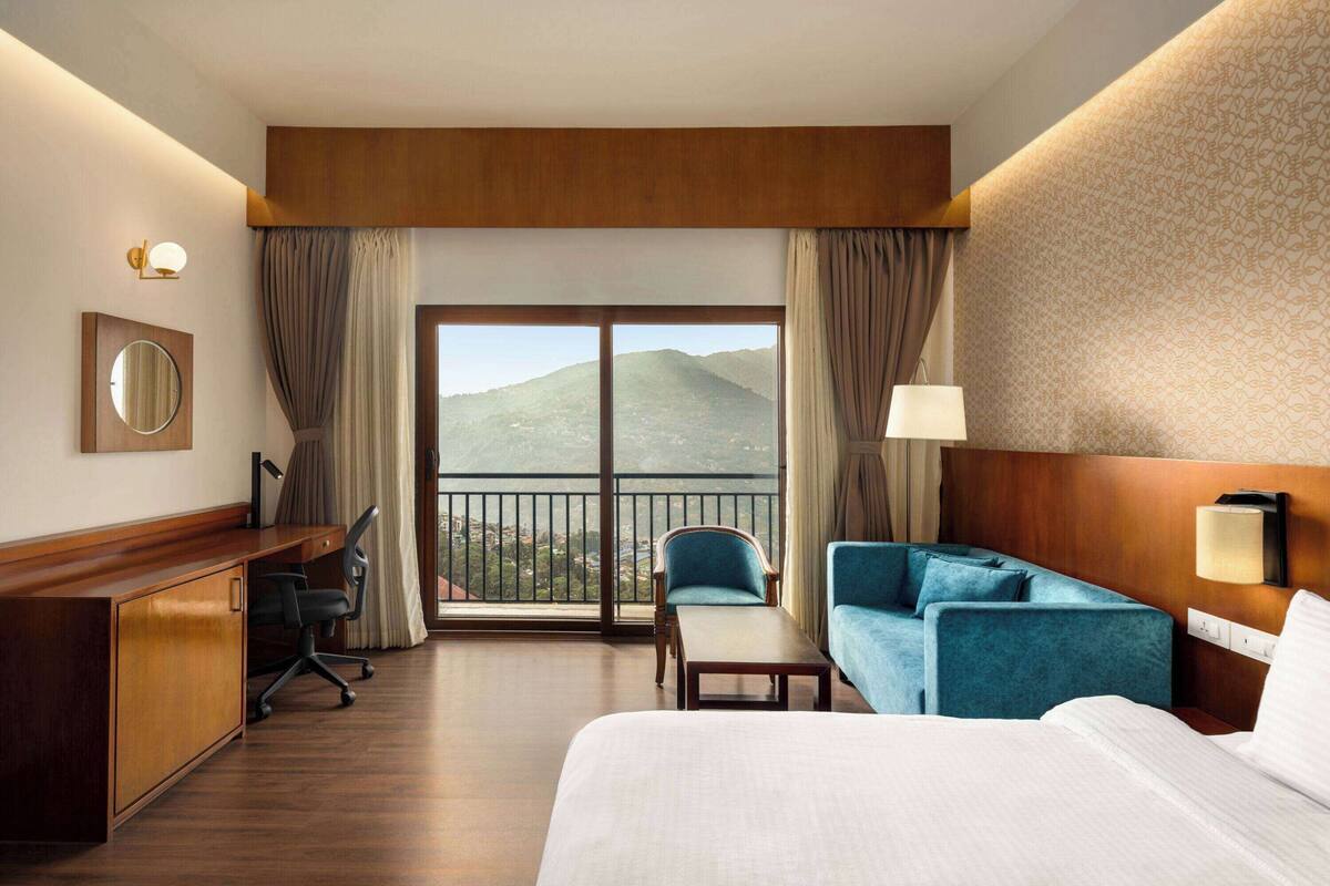 Une chambre d’hôtel comprenant un lit, un bureau, une chaise, un canapé et une vue sur les montagnes, visible à travers une grande fenêtre.
