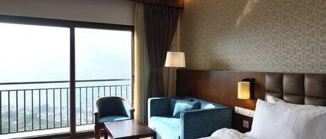 A hotel room with a large bed, a desk, two armchairs, a view of the city through large windows, and a patterned wall.