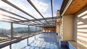 A modern indoor swimming pool with a city view, large windows, and a wooden ceiling.
