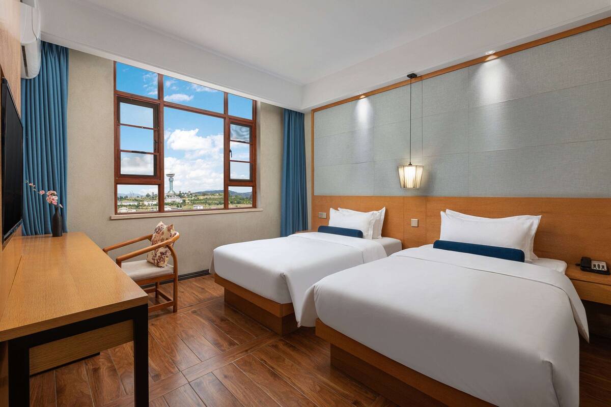 Ein Hotelzimmer mit zwei Betten, einem Holztisch, einem Stuhl, einem Fenster mit Aussicht und einer Wandlampe.