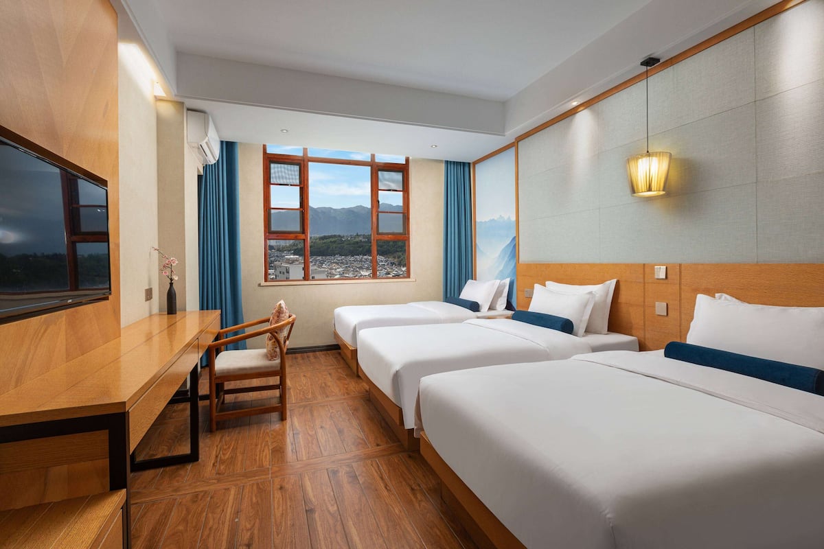 Ein Hotelzimmer mit drei Betten, einem Schreibtisch und einem Stuhl. Ein großes Fenster bietet einen Blick auf Berge und eine Stadt.