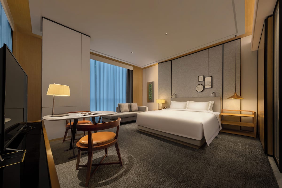 A modern hotel room with a bed, a desk with a chair, a television, and a sofa.