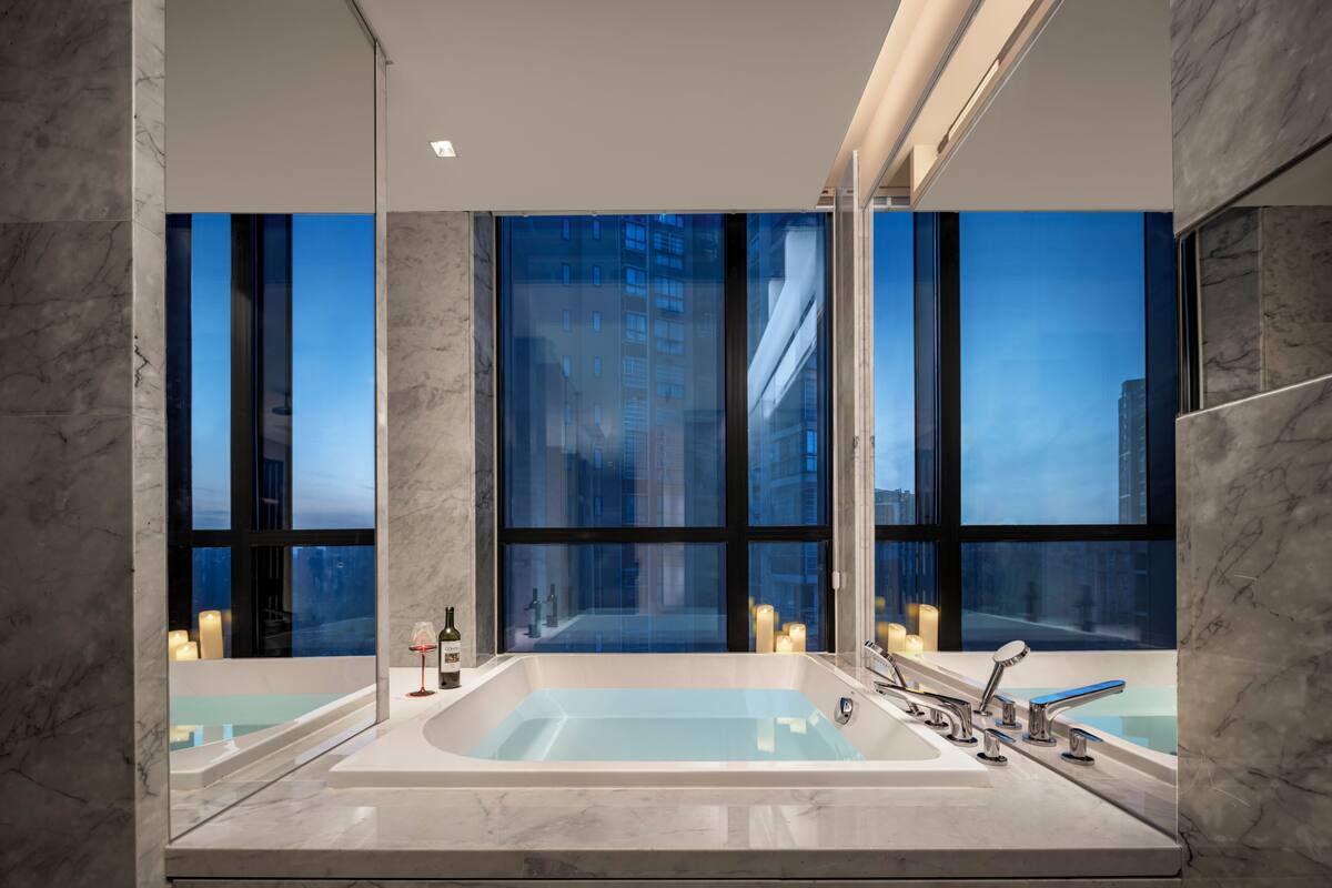 A modern bathroom with a large bathtub, two sinks, and a large window offering a city view.