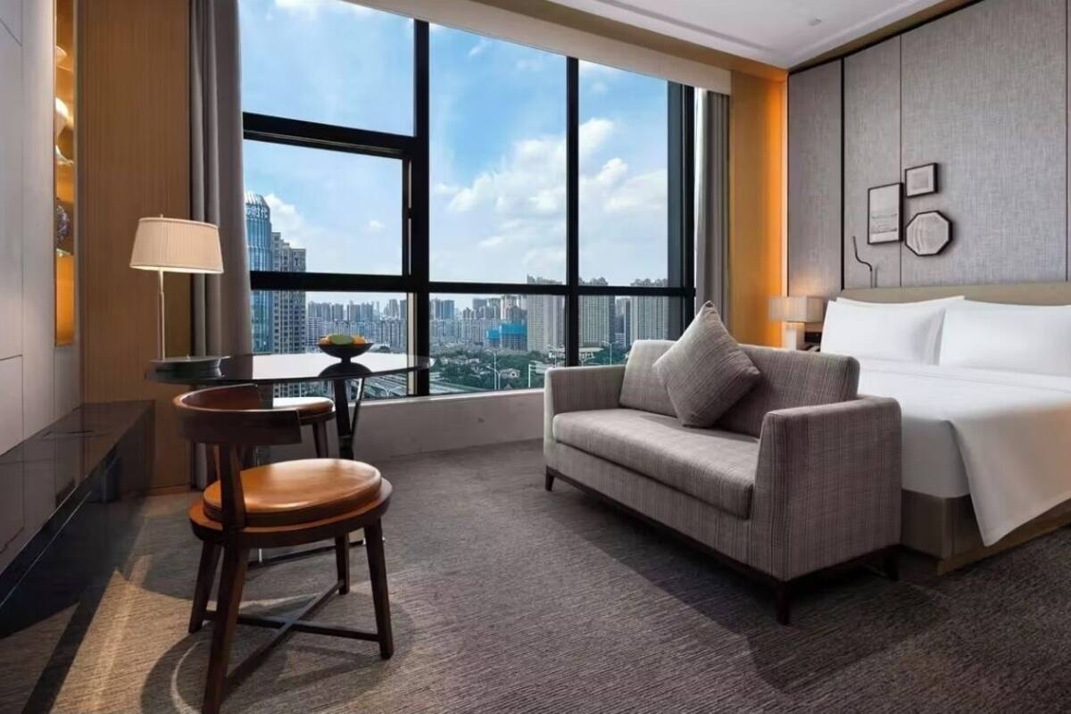 A hotel room with a large window, a sofa, a chair, a bed, and a desk with a lamp.
