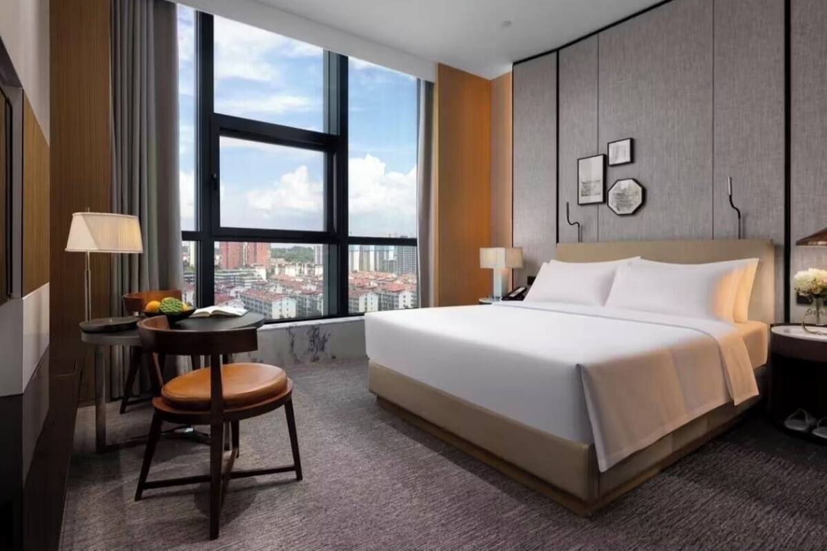 A hotel room with a large window, a bed, a desk with a chair, and a nightstand.