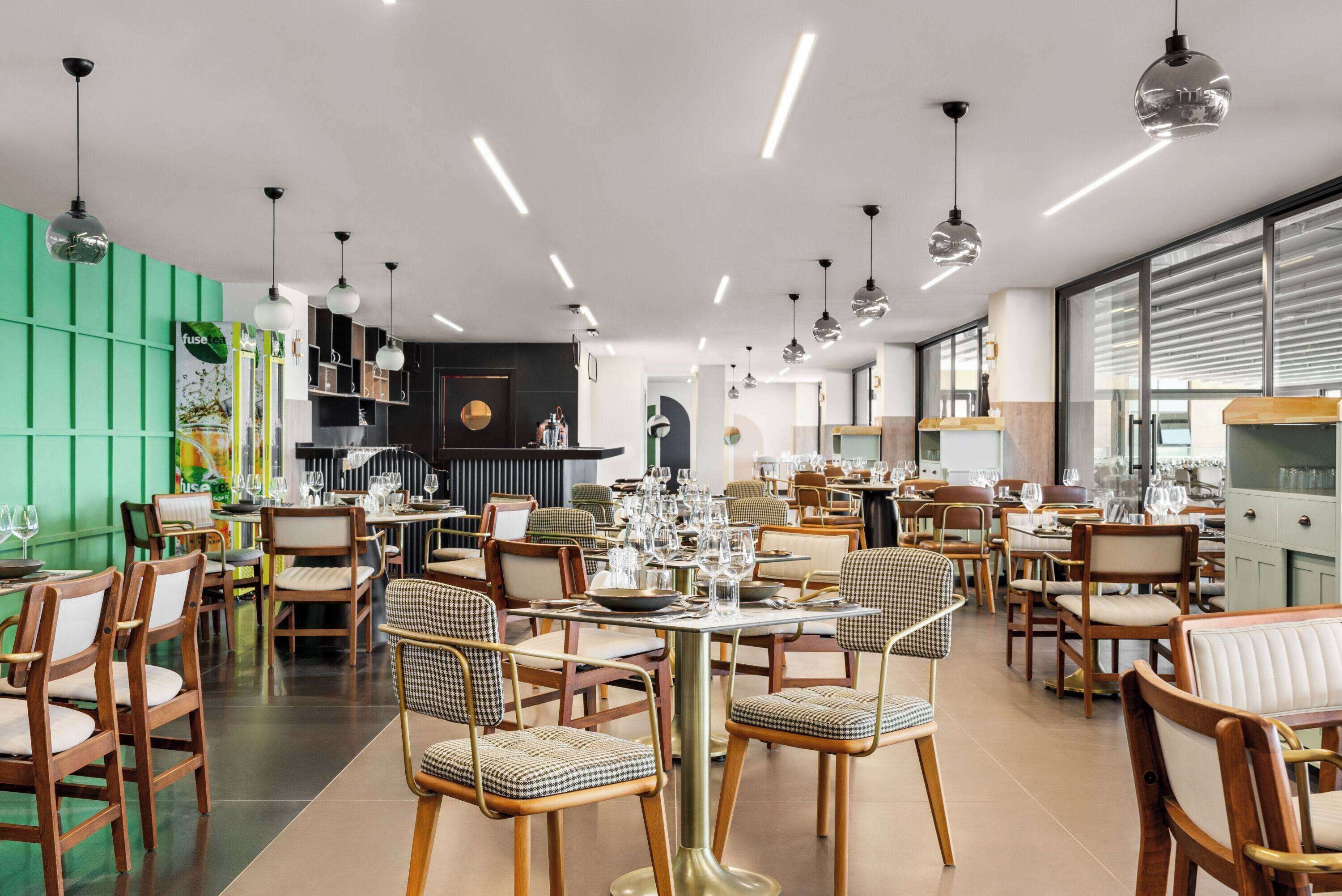 A modern restaurant with wooden chairs and tables, a green accent wall, and pendant lighting.