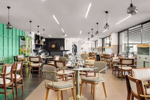 A modern restaurant with wooden chairs and tables, a green accent wall, and pendant lighting.