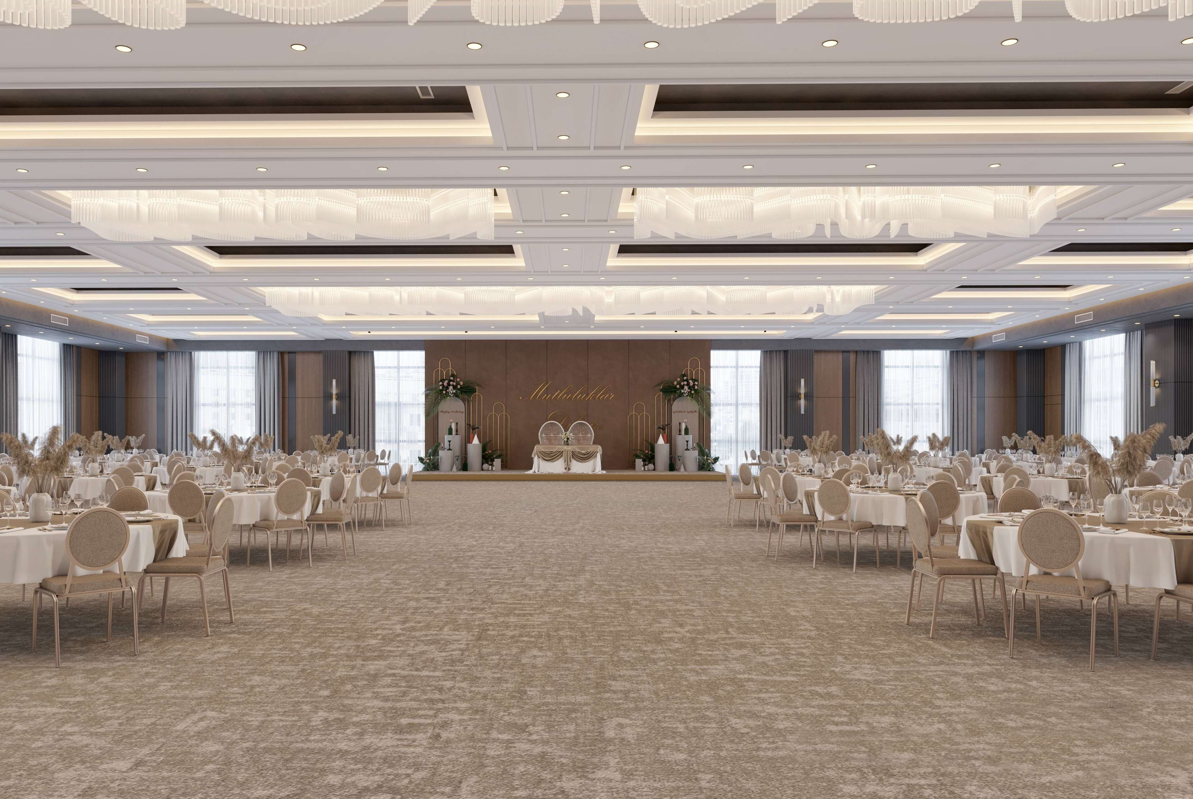 Un amplio salón de banquetes con filas de mesas y sillas, un escenario con un fondo floral y un diseño de techo moderno.