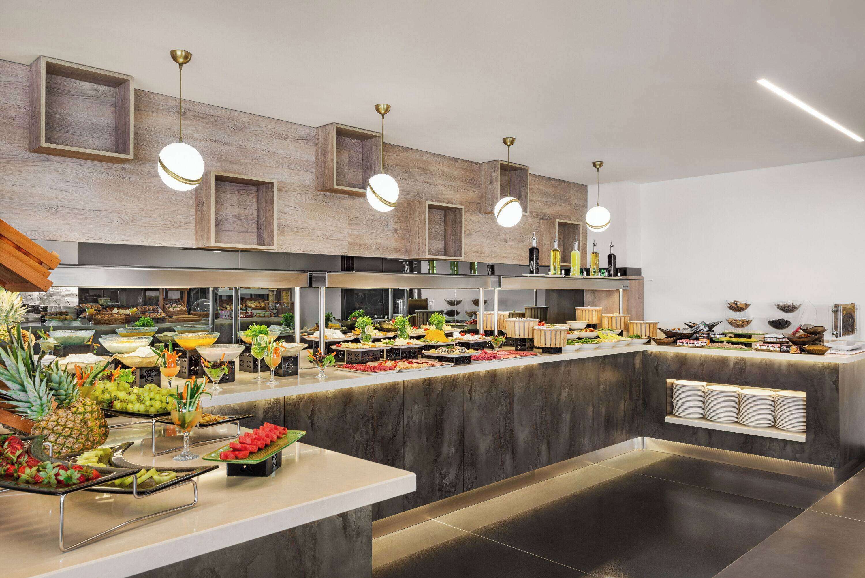 Zona de buffet con diferentes alimentos y bebidas, paneles de pared de madera e iluminación colgante.
