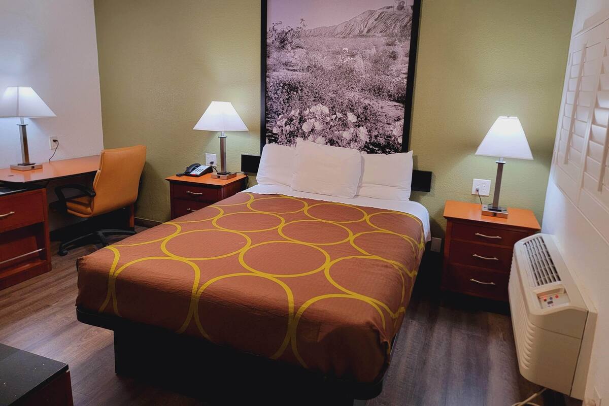 A hotel room with a bed, two bedside tables with lamps, a desk with a chair, a large framed picture on the wall, and a wall-mounted air conditioner.