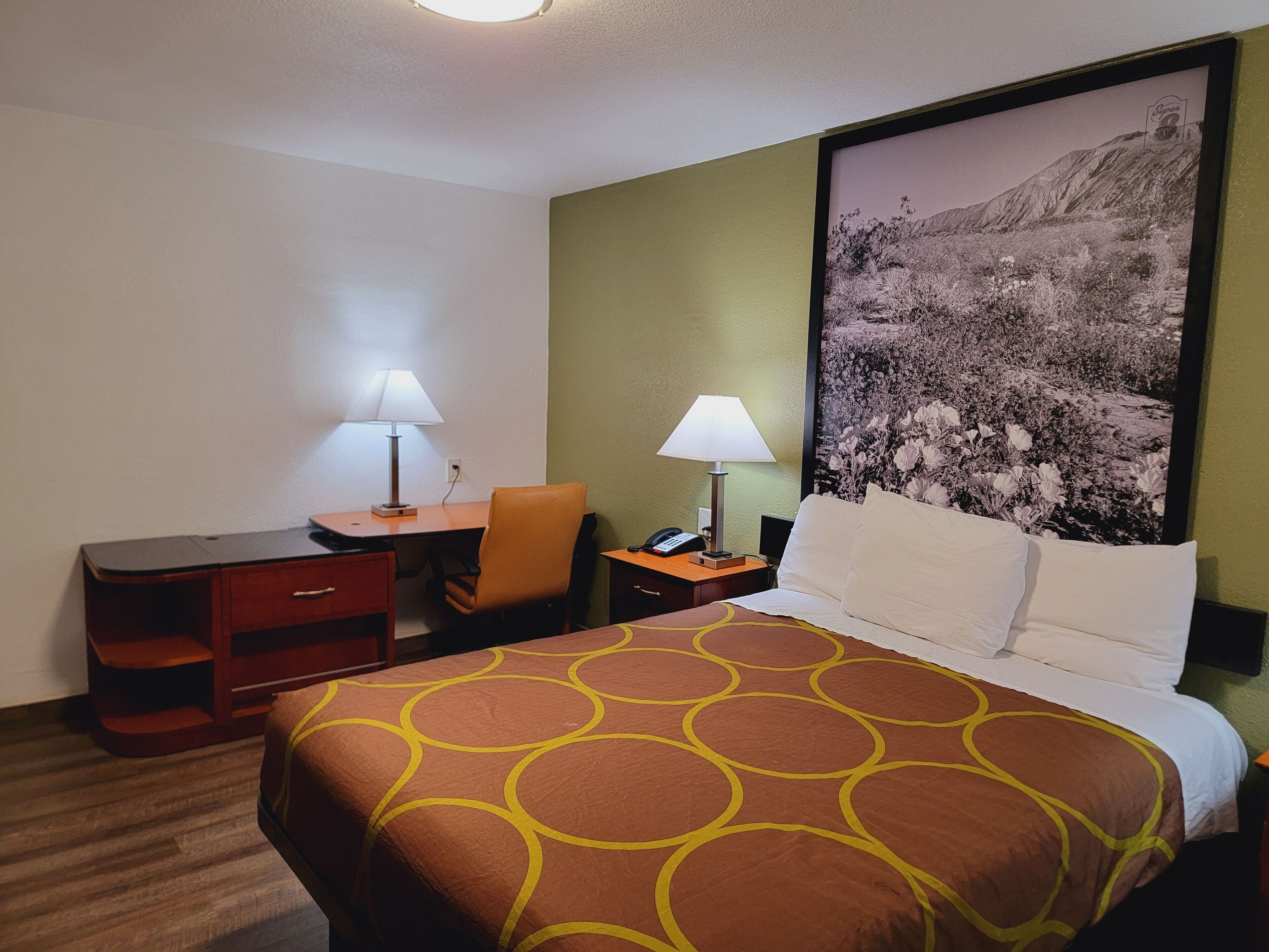 Une chambre d’hôtel avec un lit, un bureau, une chaise, des lampes et un grand tableau encadré au mur.
